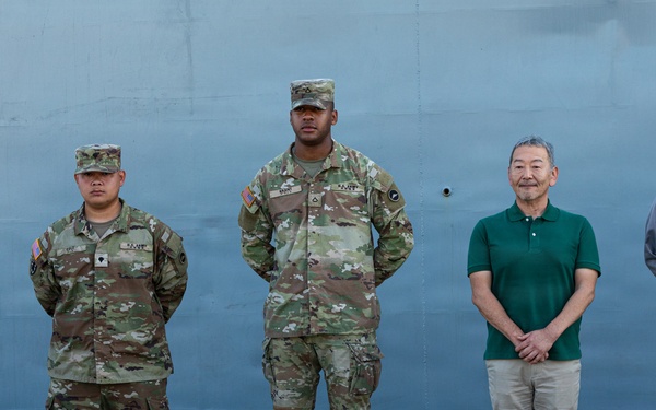 Six individuals stand in a line against a large gray metal structure, likely a ship hull or military vessel at a dock or port facility. Three individuals on the left are wearing U.S. Army OCP (Operational Camouflage Pattern) uniforms with patrol caps, while two individuals on the right are dressed in civilian clothing. The setting appears to be an official or ceremonial group photo opportunity.