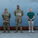Six individuals stand in a line against a large gray metal structure, likely a ship hull or military vessel at a dock or port facility. Three individuals on the left are wearing U.S. Army OCP (Operational Camouflage Pattern) uniforms with patrol caps, while two individuals on the right are dressed in civilian clothing. The setting appears to be an official or ceremonial group photo opportunity.