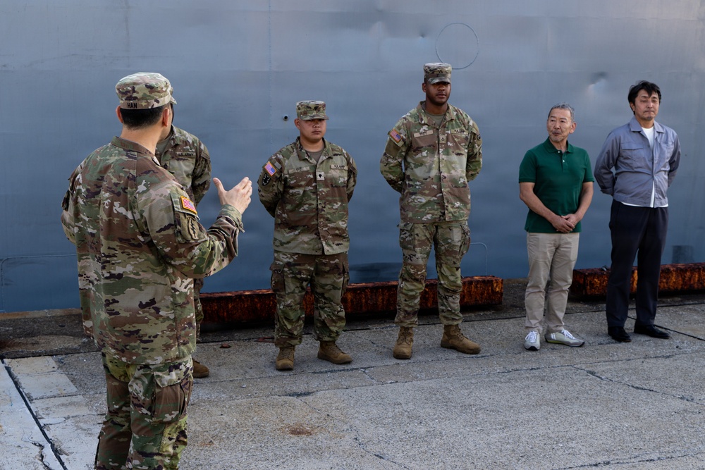 A U.S. Army soldier in OCP (Operational Camouflage Pattern) uniform is addressing a small group consisting of two other uniformed soldiers and two civilians in casual attire. The setting appears to be a military port or dock facility, with a large gray vessel hull visible in the background. The speaking soldier's name tape reads 'HAN' and the group appears to be engaged in a formal or semi-formal briefing.