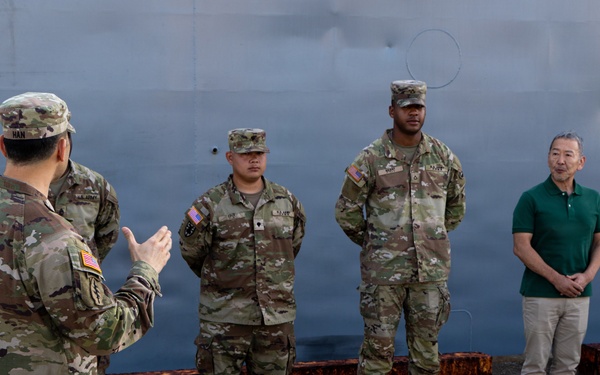 A U.S. Army soldier in OCP (Operational Camouflage Pattern) uniform is addressing a small group consisting of two other uniformed soldiers and two civilians in casual attire. The setting appears to be a military port or dock facility, with a large gray vessel hull visible in the background. The speaking soldier's name tape reads 'HAN' and the group appears to be engaged in a formal or semi-formal briefing.