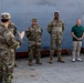 A U.S. Army soldier in OCP (Operational Camouflage Pattern) uniform is addressing a small group consisting of two other uniformed soldiers and two civilians in casual attire. The setting appears to be a military port or dock facility, with a large gray vessel hull visible in the background. The speaking soldier's name tape reads 'HAN' and the group appears to be engaged in a formal or semi-formal briefing.