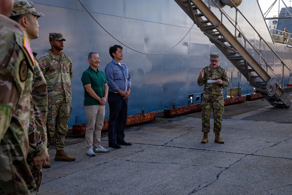 A group of U.S. Army soldiers and two civilians in casual attire are gathered at a port or dock facility beside a large vessel. A soldier on the right appears to be addressing the group while holding a document. The setting suggests an official briefing or welcoming ceremony at a maritime installation.