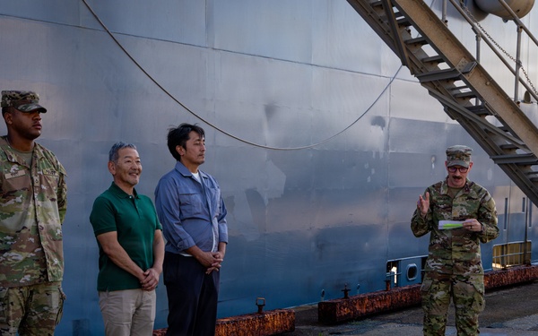 A group of U.S. Army soldiers and two civilians in casual attire are gathered at a port or dock facility beside a large vessel. A soldier on the right appears to be addressing the group while holding a document. The setting suggests an official briefing or welcoming ceremony at a maritime installation.
