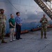 A group of U.S. Army soldiers and two civilians in casual attire are gathered at a port or dock facility beside a large vessel. A soldier on the right appears to be addressing the group while holding a document. The setting suggests an official briefing or welcoming ceremony at a maritime installation.