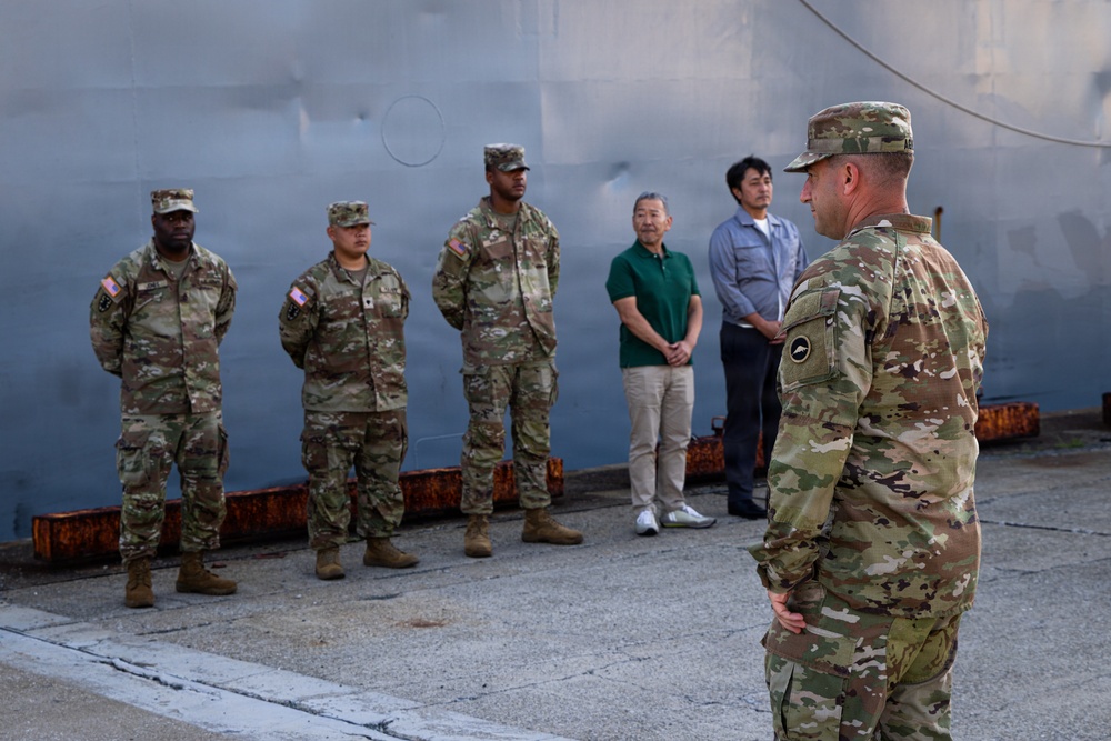 A U.S. Army officer addresses a small group consisting of three soldiers in OCP (Operational Camouflage Pattern) uniforms and two civilians in casual attire, standing in front of a large gray metal structure, likely a ship or industrial vessel. The setting appears to be a port or maritime facility, suggested by the rusted dock infrastructure visible in the foreground. The formation suggests a briefing or formal address.