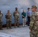 A U.S. Army officer addresses a small group consisting of three soldiers in OCP (Operational Camouflage Pattern) uniforms and two civilians in casual attire, standing in front of a large gray metal structure, likely a ship or industrial vessel. The setting appears to be a port or maritime facility, suggested by the rusted dock infrastructure visible in the foreground. The formation suggests a briefing or formal address.