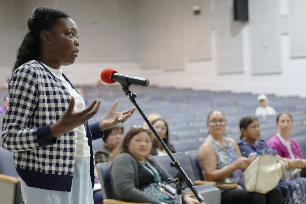 A woman is standing at a microphone stand with a red windscreen in what appears to be an auditorium or community meeting hall, speaking to an audience. She is wearing a navy and white checkered cardigan over a white outfit and is gesturing with both hands while speaking. Several audience members are seated in blue auditorium chairs in the background, attentively observing.