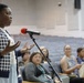 A woman is standing at a microphone stand with a red windscreen in what appears to be an auditorium or community meeting hall, speaking to an audience. She is wearing a navy and white checkered cardigan over a white outfit and is gesturing with both hands while speaking. Several audience members are seated in blue auditorium chairs in the background, attentively observing.