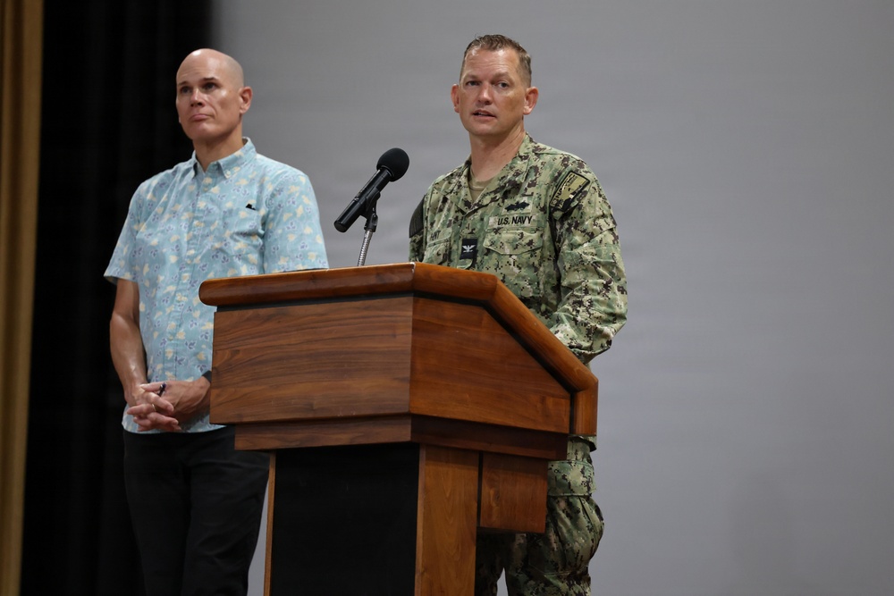 A U.S. Navy officer in digital camouflage uniform is speaking at a wooden podium with a microphone. A civilian man in a light blue floral/patterned button-up shirt stands behind and to the left, observing. The setting appears to be an indoor auditorium or briefing room with a neutral gray backdrop.