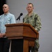 A U.S. Navy officer in digital camouflage uniform is speaking at a wooden podium with a microphone. A civilian man in a light blue floral/patterned button-up shirt stands behind and to the left, observing. The setting appears to be an indoor auditorium or briefing room with a neutral gray backdrop.