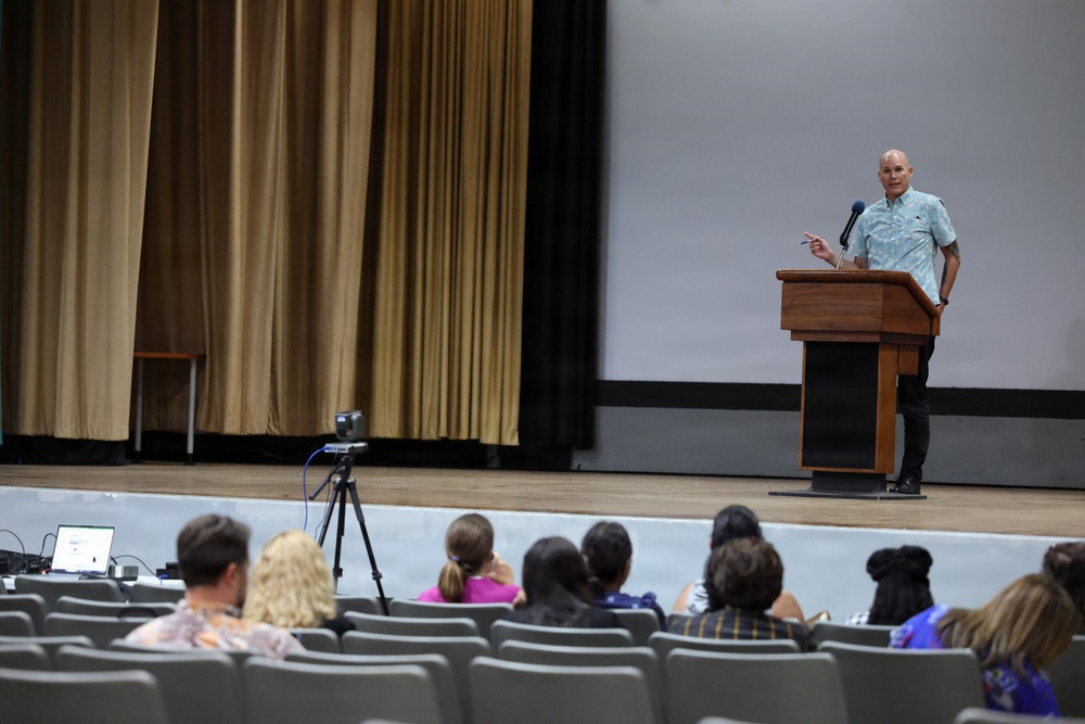A bald male speaker stands at a wooden podium on an auditorium stage, addressing a seated audience. A projection screen is visible behind him, and a video camera on a tripod is positioned in front of the stage to record the event. The audience occupies several rows of gray theater-style seats.