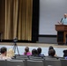 A bald male speaker stands at a wooden podium on an auditorium stage, addressing a seated audience. A projection screen is visible behind him, and a video camera on a tripod is positioned in front of the stage to record the event. The audience occupies several rows of gray theater-style seats.
