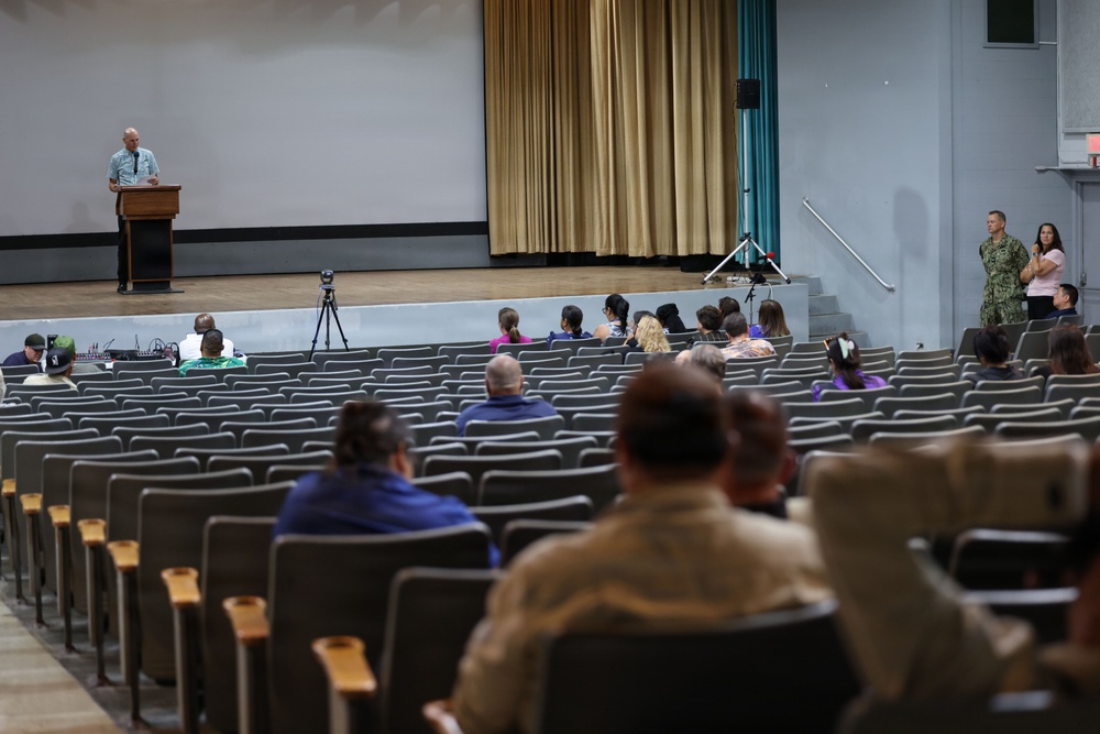 A sparsely attended presentation or briefing is taking place in a large auditorium or theater. A speaker stands at a podium on the elevated stage while a small audience is seated in the mostly empty rows of chairs. At least one person in military uniform is visible near the right side of the room.