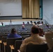 A sparsely attended presentation or briefing is taking place in a large auditorium or theater. A speaker stands at a podium on the elevated stage while a small audience is seated in the mostly empty rows of chairs. At least one person in military uniform is visible near the right side of the room.