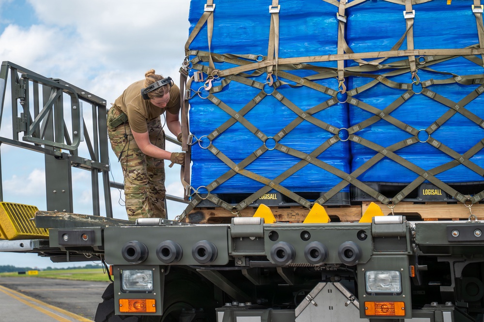 A military service member in OCP (Operational Camouflage Pattern) uniform is securing cargo straps on a military flatbed vehicle loaded with blue-wrapped pallets. The pallets are secured with a military-standard cargo net featuring D-rings and carabiner clips. The setting appears to be an airfield or military installation, suggesting air cargo loading or transport operations.