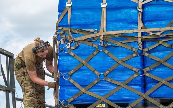A military service member in OCP (Operational Camouflage Pattern) uniform is securing cargo straps on a military flatbed vehicle loaded with blue-wrapped pallets. The pallets are secured with a military-standard cargo net featuring D-rings and carabiner clips. The setting appears to be an airfield or military installation, suggesting air cargo loading or transport operations.