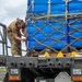 A military service member in OCP (Operational Camouflage Pattern) uniform is securing cargo straps on a military flatbed vehicle loaded with blue-wrapped pallets. The pallets are secured with a military-standard cargo net featuring D-rings and carabiner clips. The setting appears to be an airfield or military installation, suggesting air cargo loading or transport operations.