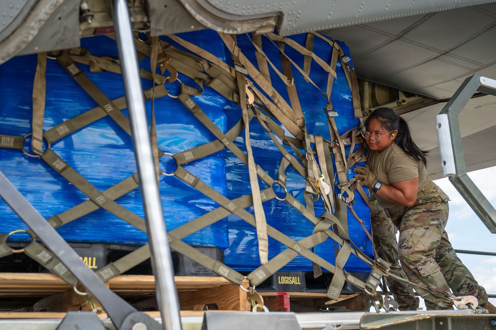 A military service member in OCP (Operational Camouflage Pattern) uniform is loading or securing a large cargo pallet wrapped in blue tarpaulin onto what appears to be a military transport aircraft. The pallet is secured with a tan cargo net and restraint straps, and equipment labeled 'LOGISALL' is visible on the loading platform. The scene depicts standard military aerial cargo loading operations.