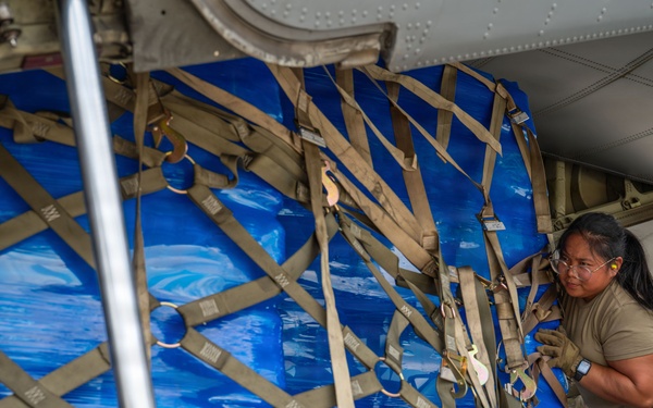 A military service member in OCP (Operational Camouflage Pattern) uniform is loading or securing a large cargo pallet wrapped in blue tarpaulin onto what appears to be a military transport aircraft. The pallet is secured with a tan cargo net and restraint straps, and equipment labeled 'LOGISALL' is visible on the loading platform. The scene depicts standard military aerial cargo loading operations.