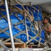 A military service member in OCP (Operational Camouflage Pattern) uniform is loading or securing a large cargo pallet wrapped in blue tarpaulin onto what appears to be a military transport aircraft. The pallet is secured with a tan cargo net and restraint straps, and equipment labeled 'LOGISALL' is visible on the loading platform. The scene depicts standard military aerial cargo loading operations.