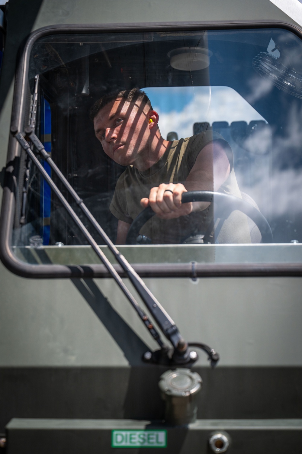 A young male in a olive/green sleeveless military-style shirt is seated in the cab of a large diesel-powered military or heavy-duty vehicle, gripping the steering wheel. He is wearing yellow ear protection (earplugs) and looking outward through the windshield. The vehicle exterior is painted olive drab and bears a green 'DIESEL' fuel label.