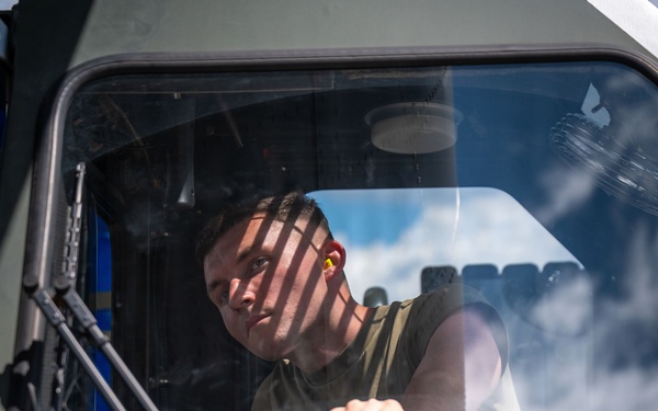 A young male in a olive/green sleeveless military-style shirt is seated in the cab of a large diesel-powered military or heavy-duty vehicle, gripping the steering wheel. He is wearing yellow ear protection (earplugs) and looking outward through the windshield. The vehicle exterior is painted olive drab and bears a green 'DIESEL' fuel label.