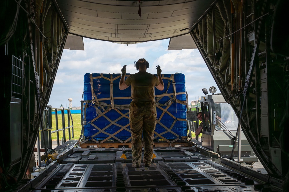 A military loadmaster in camouflage uniform stands at the open rear ramp of a military cargo aircraft, using hand signals to guide the loading of a large blue-wrapped pallet. The pallet is secured with tan cargo straps in a cross-hatch pattern and is being moved by a cargo loader vehicle operated by a ground crew member. The scene appears to take place on a military airfield under partly cloudy skies.
