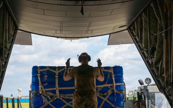 A military loadmaster in camouflage uniform stands at the open rear ramp of a military cargo aircraft, using hand signals to guide the loading of a large blue-wrapped pallet. The pallet is secured with tan cargo straps in a cross-hatch pattern and is being moved by a cargo loader vehicle operated by a ground crew member. The scene appears to take place on a military airfield under partly cloudy skies.