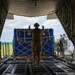 A military loadmaster in camouflage uniform stands at the open rear ramp of a military cargo aircraft, using hand signals to guide the loading of a large blue-wrapped pallet. The pallet is secured with tan cargo straps in a cross-hatch pattern and is being moved by a cargo loader vehicle operated by a ground crew member. The scene appears to take place on a military airfield under partly cloudy skies.