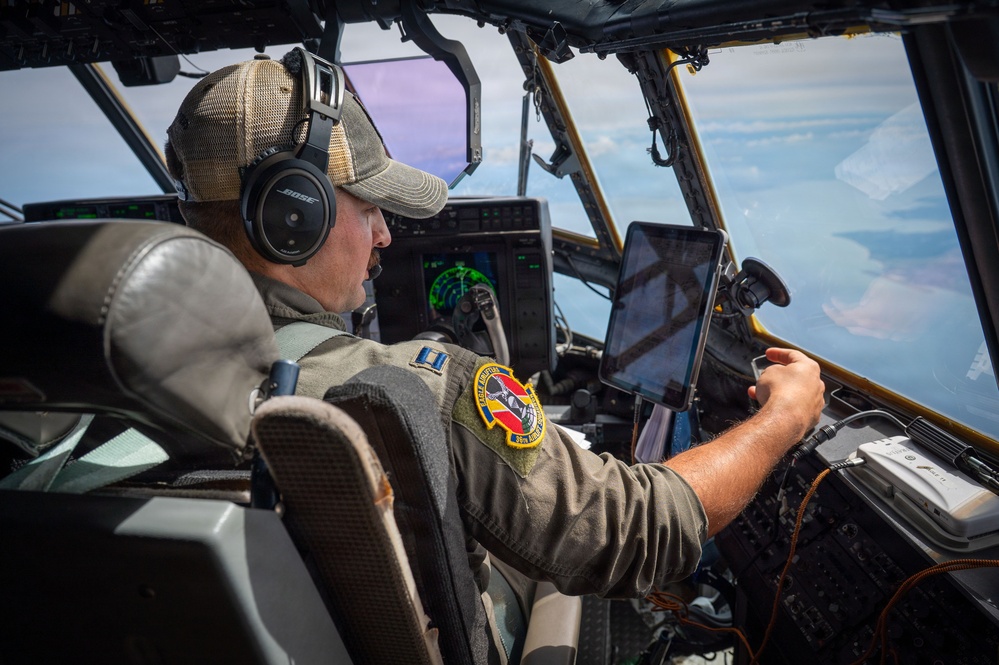 A military pilot is seated in the cockpit of a large aircraft, actively operating the controls while in flight. He is wearing a flight suit with a unit patch, a baseball cap, and Bose aviation headphones. A tablet device is mounted on the windscreen frame, and aerial terrain is visible through the cockpit windows.