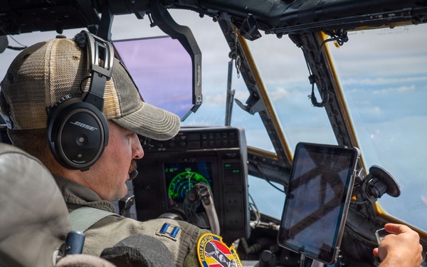 A military pilot is seated in the cockpit of a large aircraft, actively operating the controls while in flight. He is wearing a flight suit with a unit patch, a baseball cap, and Bose aviation headphones. A tablet device is mounted on the windscreen frame, and aerial terrain is visible through the cockpit windows.