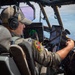 A military pilot is seated in the cockpit of a large aircraft, actively operating the controls while in flight. He is wearing a flight suit with a unit patch, a baseball cap, and Bose aviation headphones. A tablet device is mounted on the windscreen frame, and aerial terrain is visible through the cockpit windows.