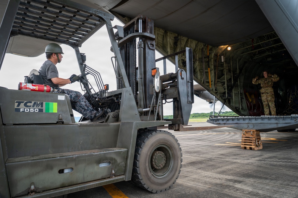 A military loading operation is underway on an airfield tarmac, where a TCM FD50 forklift operated by a helmeted individual in military-style camouflage pants is approaching the open rear cargo ramp of a large military transport aircraft. A soldier in full camouflage uniform stands inside the aircraft's cargo bay, appearing to direct or guide the forklift operator. The scene suggests a joint or multinational military logistics exercise or operation.