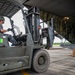 A military loading operation is underway on an airfield tarmac, where a TCM FD50 forklift operated by a helmeted individual in military-style camouflage pants is approaching the open rear cargo ramp of a large military transport aircraft. A soldier in full camouflage uniform stands inside the aircraft's cargo bay, appearing to direct or guide the forklift operator. The scene suggests a joint or multinational military logistics exercise or operation.
