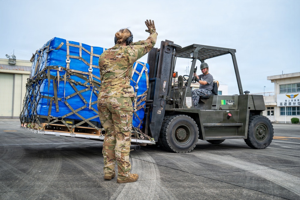 A military service member in OCP (Operational Camouflage Pattern) uniform is directing a forklift operator who is moving a large cargo pallet wrapped in blue tarpaulin and secured with cargo netting and straps. The operation appears to be taking place on a military airfield tarmac, with Japanese text visible on a building in the background, suggesting a location in Japan. This appears to be a cargo offload or logistics operation at a joint military installation.