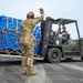 A military service member in OCP (Operational Camouflage Pattern) uniform is directing a forklift operator who is moving a large cargo pallet wrapped in blue tarpaulin and secured with cargo netting and straps. The operation appears to be taking place on a military airfield tarmac, with Japanese text visible on a building in the background, suggesting a location in Japan. This appears to be a cargo offload or logistics operation at a joint military installation.