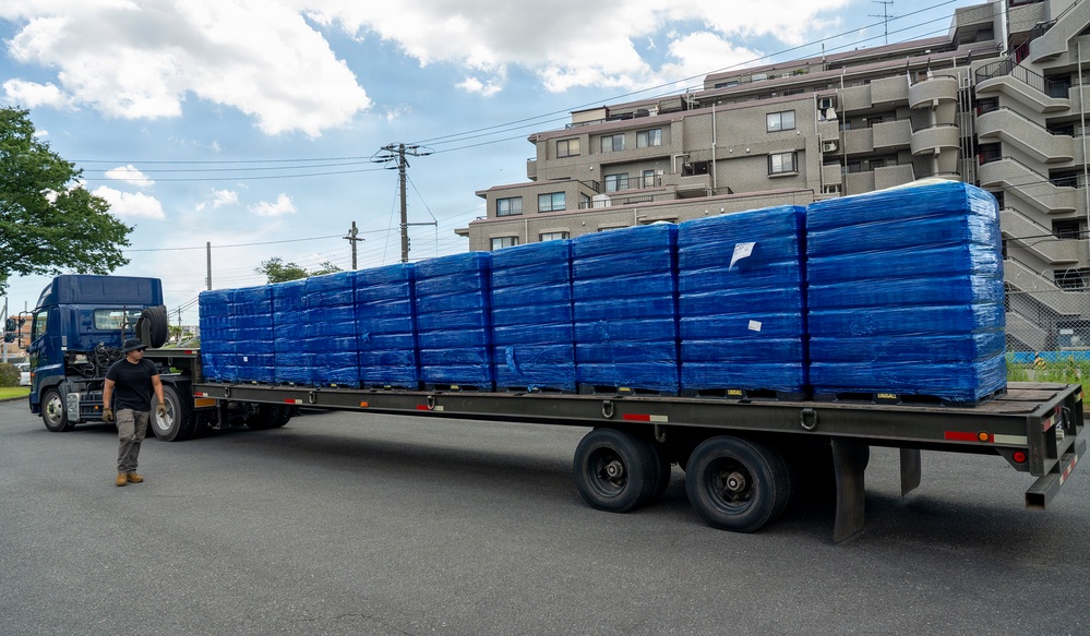 A flatbed semi-truck is loaded with numerous pallets wrapped in blue stretch film, parked in what appears to be a Japanese urban area. A single worker in dark clothing stands beside the truck, likely inspecting or overseeing the cargo. A multi-story residential building and utility poles are visible in the background.