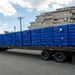 A flatbed semi-truck is loaded with numerous pallets wrapped in blue stretch film, parked in what appears to be a Japanese urban area. A single worker in dark clothing stands beside the truck, likely inspecting or overseeing the cargo. A multi-story residential building and utility poles are visible in the background.