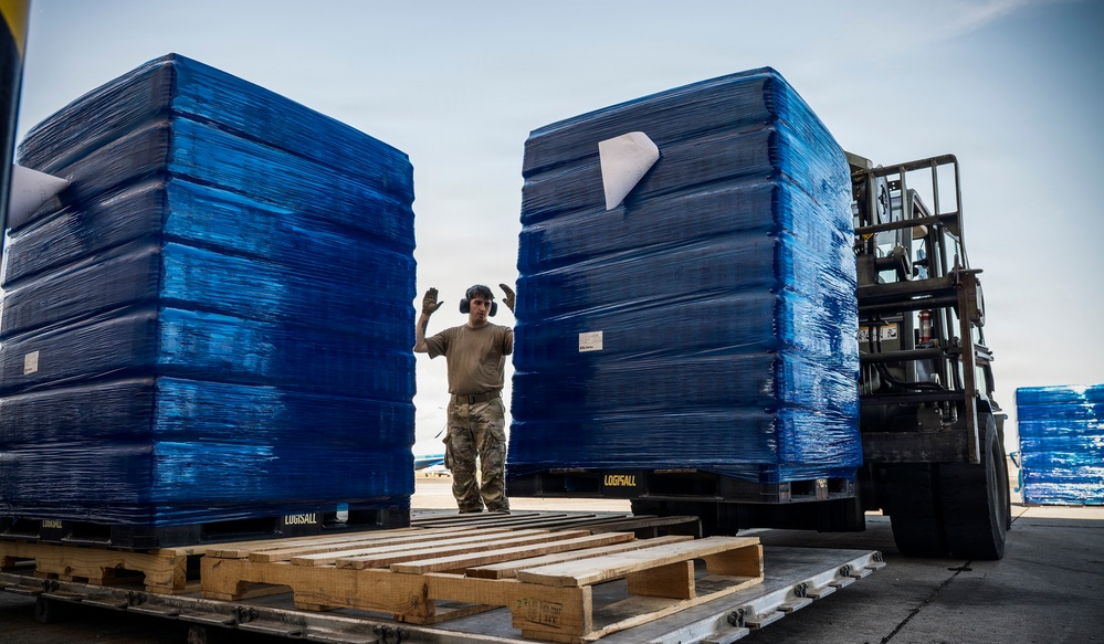 A military service member in combat uniform is directing a forklift operator handling large pallets of blue shrink-wrapped cargo on what appears to be a military airfield or logistics staging area. The worker is using hand signals to guide the placement of the heavy pallets, which are stacked on wooden LOGFSALL-branded military pallets. The scene depicts a standard military cargo loading or unloading operation.