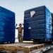 A military service member in combat uniform is directing a forklift operator handling large pallets of blue shrink-wrapped cargo on what appears to be a military airfield or logistics staging area. The worker is using hand signals to guide the placement of the heavy pallets, which are stacked on wooden LOGFSALL-branded military pallets. The scene depicts a standard military cargo loading or unloading operation.
