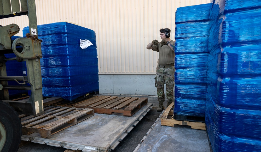 A military service member in camouflage uniform and hearing protection is working in a loading dock area surrounded by large pallets wrapped in blue stretch film. A forklift is visible on the left side, and the pallets appear to be staged for loading or unloading operations. The setting appears to be a military logistics or supply facility.