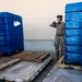 A military service member in camouflage uniform and hearing protection is working in a loading dock area surrounded by large pallets wrapped in blue stretch film. A forklift is visible on the left side, and the pallets appear to be staged for loading or unloading operations. The setting appears to be a military logistics or supply facility.