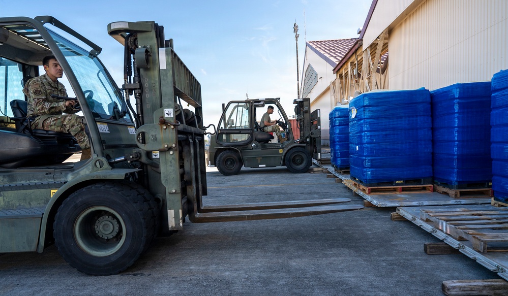 Two military personnel in camouflage uniforms are operating olive-drab forklifts in an outdoor logistics area adjacent to a building. They appear to be maneuvering to handle palletized cargo wrapped in blue plastic shrink wrap. The scene suggests a military cargo handling or humanitarian aid distribution operation.