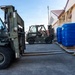 Two military personnel in camouflage uniforms are operating olive-drab forklifts in an outdoor logistics area adjacent to a building. They appear to be maneuvering to handle palletized cargo wrapped in blue plastic shrink wrap. The scene suggests a military cargo handling or humanitarian aid distribution operation.