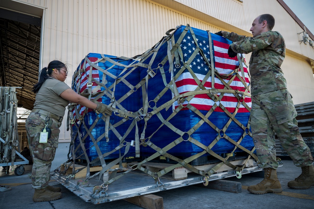 Two U.S. military personnel in OCP (Operational Camouflage Pattern) uniforms are handling a large cargo pallet wrapped in blue plastic sheeting and covered with multiple American flags, secured with cargo netting and straps. The pallet appears to be on a metal aircraft cargo loader or dolly outside a military hangar or warehouse facility. This type of configuration is consistent with dignified transfer or ceremonial cargo handling procedures.