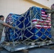Two U.S. military personnel in OCP (Operational Camouflage Pattern) uniforms are handling a large cargo pallet wrapped in blue plastic sheeting and covered with multiple American flags, secured with cargo netting and straps. The pallet appears to be on a metal aircraft cargo loader or dolly outside a military hangar or warehouse facility. This type of configuration is consistent with dignified transfer or ceremonial cargo handling procedures.