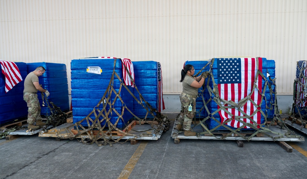 Two military personnel in camouflage uniforms are securing cargo pallets wrapped in blue plastic and draped with American flags using cargo straps and netting. The pallets are positioned on metal aircraft cargo platforms outside what appears to be a military hangar or warehouse. The scene suggests preparation for air transport of military cargo.