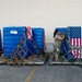 Two military personnel in camouflage uniforms are securing cargo pallets wrapped in blue plastic and draped with American flags using cargo straps and netting. The pallets are positioned on metal aircraft cargo platforms outside what appears to be a military hangar or warehouse. The scene suggests preparation for air transport of military cargo.