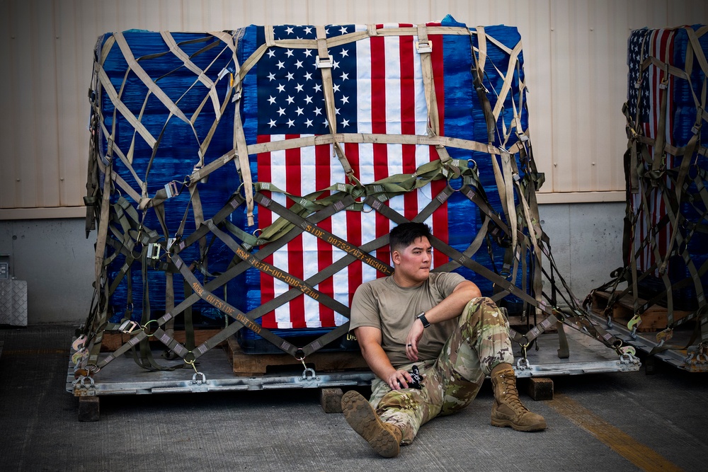 A military service member in U.S. Army/Air Force multicam (OCP) uniform sits on a cargo pallet in what appears to be a hangar or cargo facility. Behind the individual is a large military cargo net-secured pallet wrapped in blue tarpaulin with an American flag displayed prominently on its face. The scene suggests a logistics or airlift operation, possibly during a deployment or redeployment.