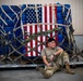 A military service member in U.S. Army/Air Force multicam (OCP) uniform sits on a cargo pallet in what appears to be a hangar or cargo facility. Behind the individual is a large military cargo net-secured pallet wrapped in blue tarpaulin with an American flag displayed prominently on its face. The scene suggests a logistics or airlift operation, possibly during a deployment or redeployment.